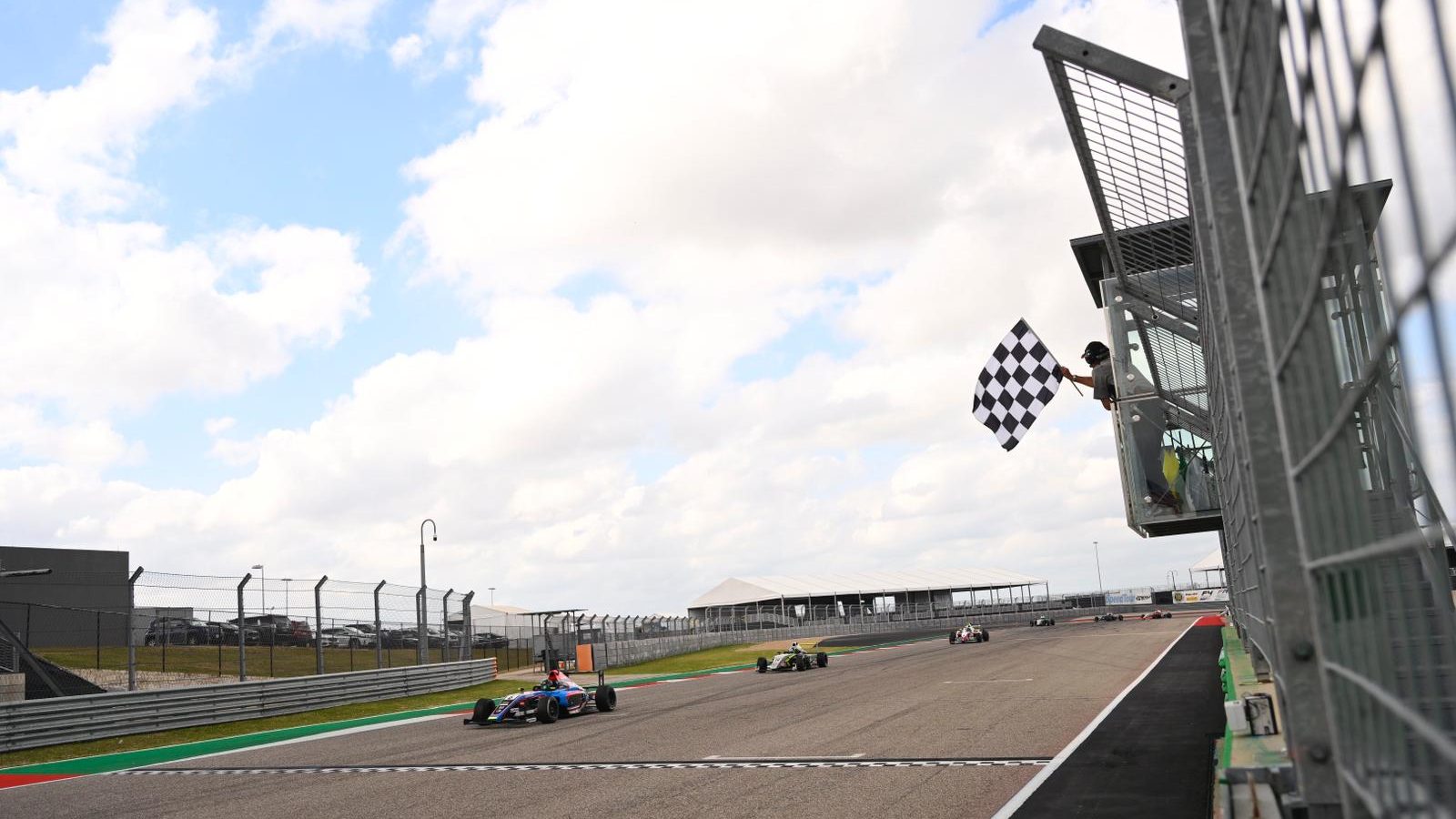 The chequered flag waves from a flag stand as cars exit from a left turn and prepare to cross a black-and-white chequered strip on the racetrack surface