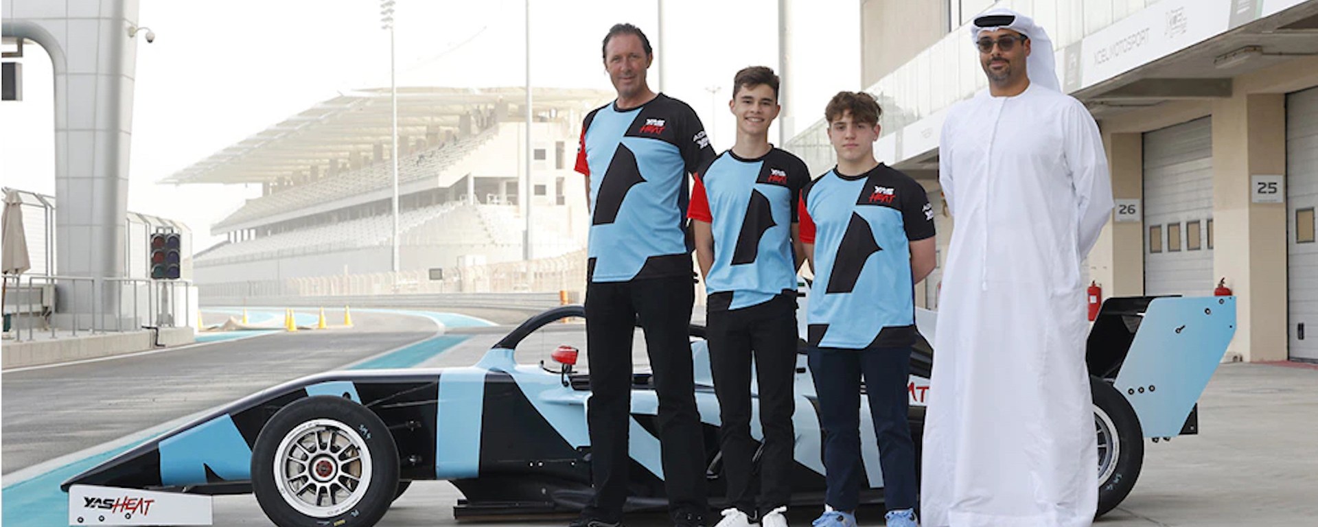Three males stand in front of a blue and black racecar wearing blue, black and red Yas Heat–branded t-shirts. A man in a thoub and keffiyeh stands beside them.