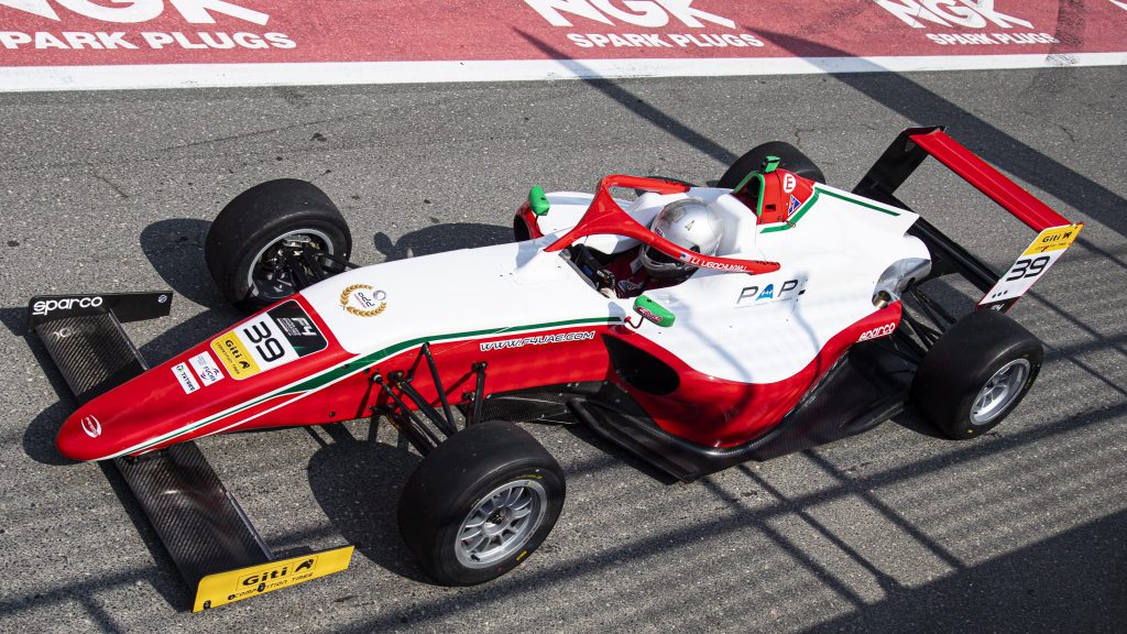 The red and white car of Ugo Ugochukwu strolling down pit lane past an NGK spark plugs sign, photographed from above left