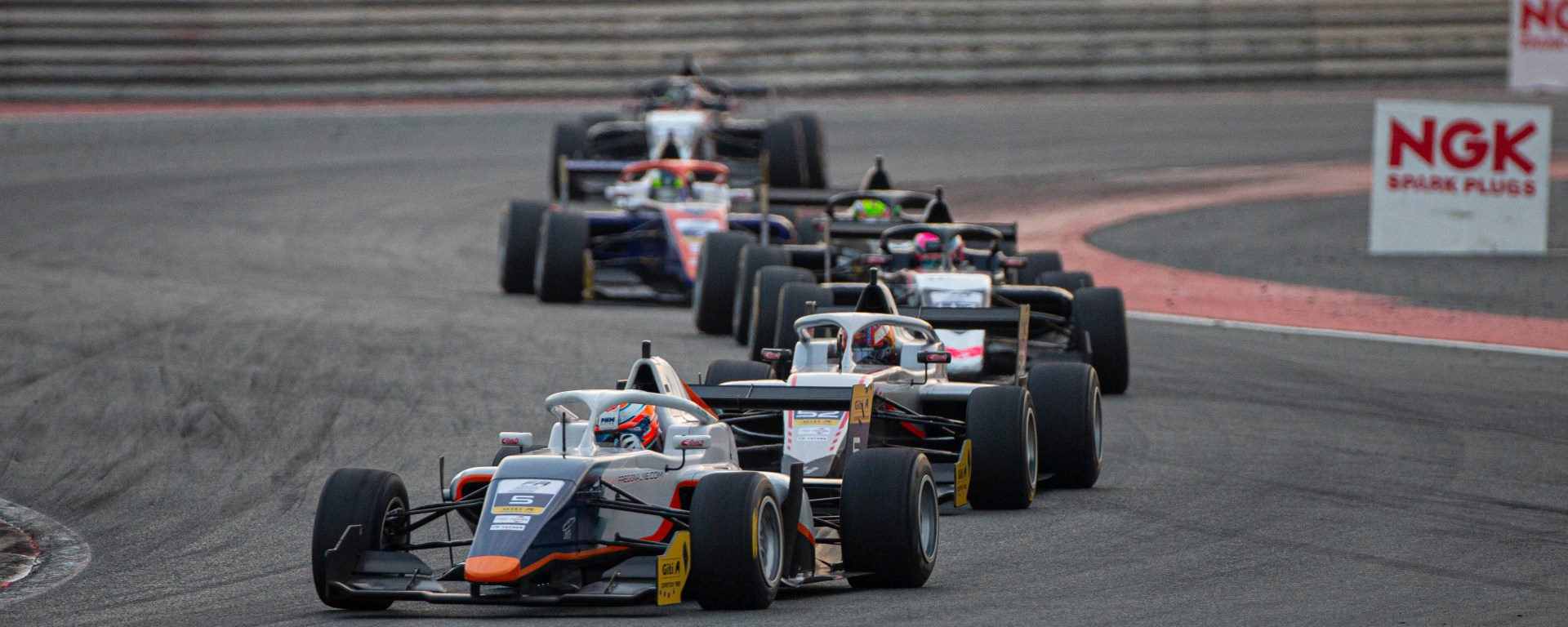 A blue and white car with orange accents leads a train of five cars navigating a left-right corner complex with NGK Spark Plugs bollards in the background