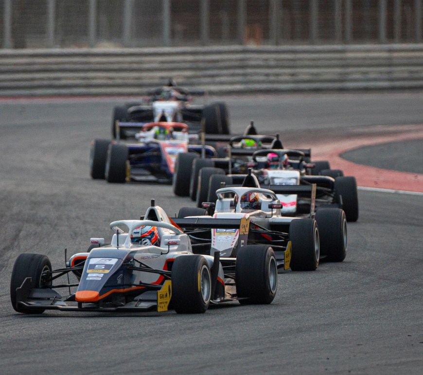 A blue and white car with orange accents leads a train of five cars navigating a left-right corner complex with NGK Spark Plugs bollards in the background