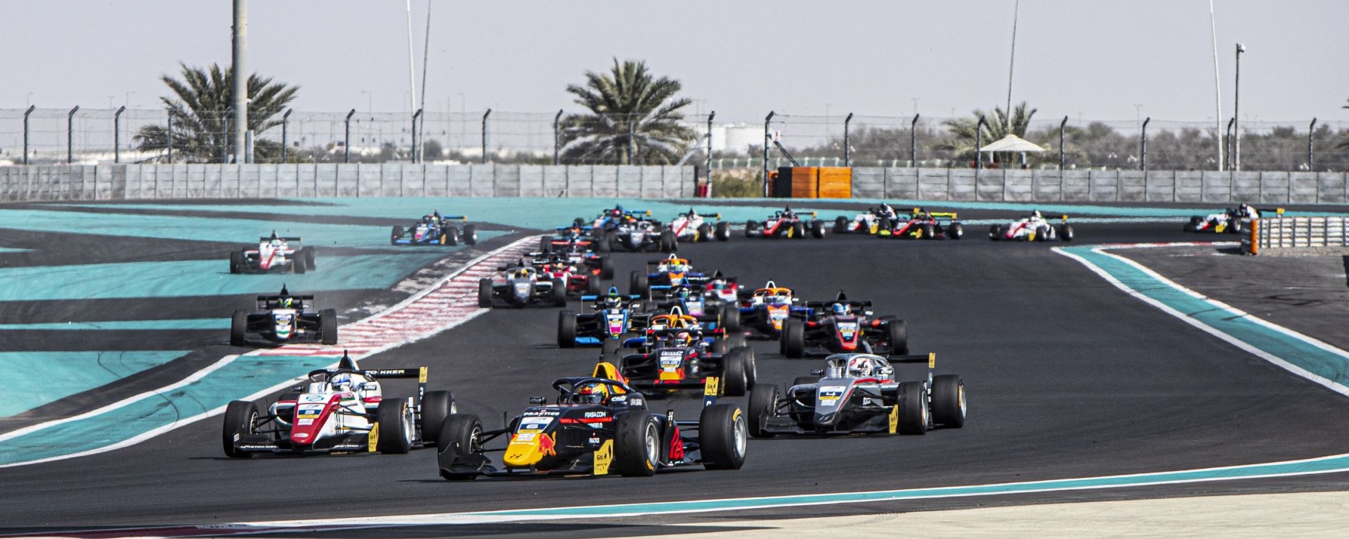 Cars navigate a slight right turn on the Yas Marina Circuit, with the characteristic teal and grey asphalt run-off traversed by three cars in the background
