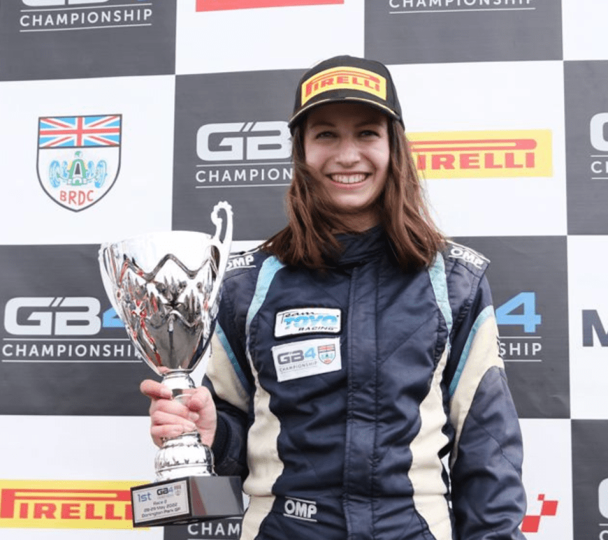 A woman (Megan Gilkes) wearing a black Pirelli cap and navy racing overalls stands on a podium holds a trophy in her right hand. The trophy's base reads '1st, Race 2, 28–29 May 2022, Donington Park GP'