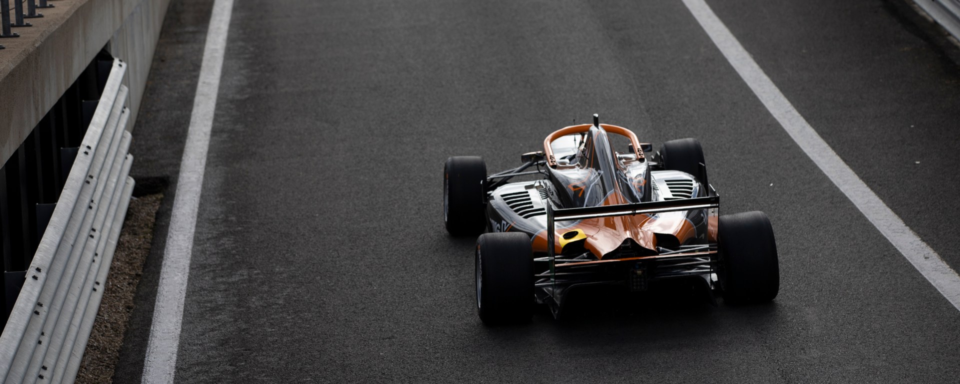 A close-up of a black and orange car leaving the pit lane at Silverstone, viewed from above rear