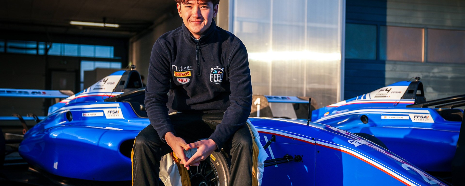 Kevin Foster, wearing a FEED Racing jacket, sits on the right-front tyre of a blue single-seater and smiles at the camera.