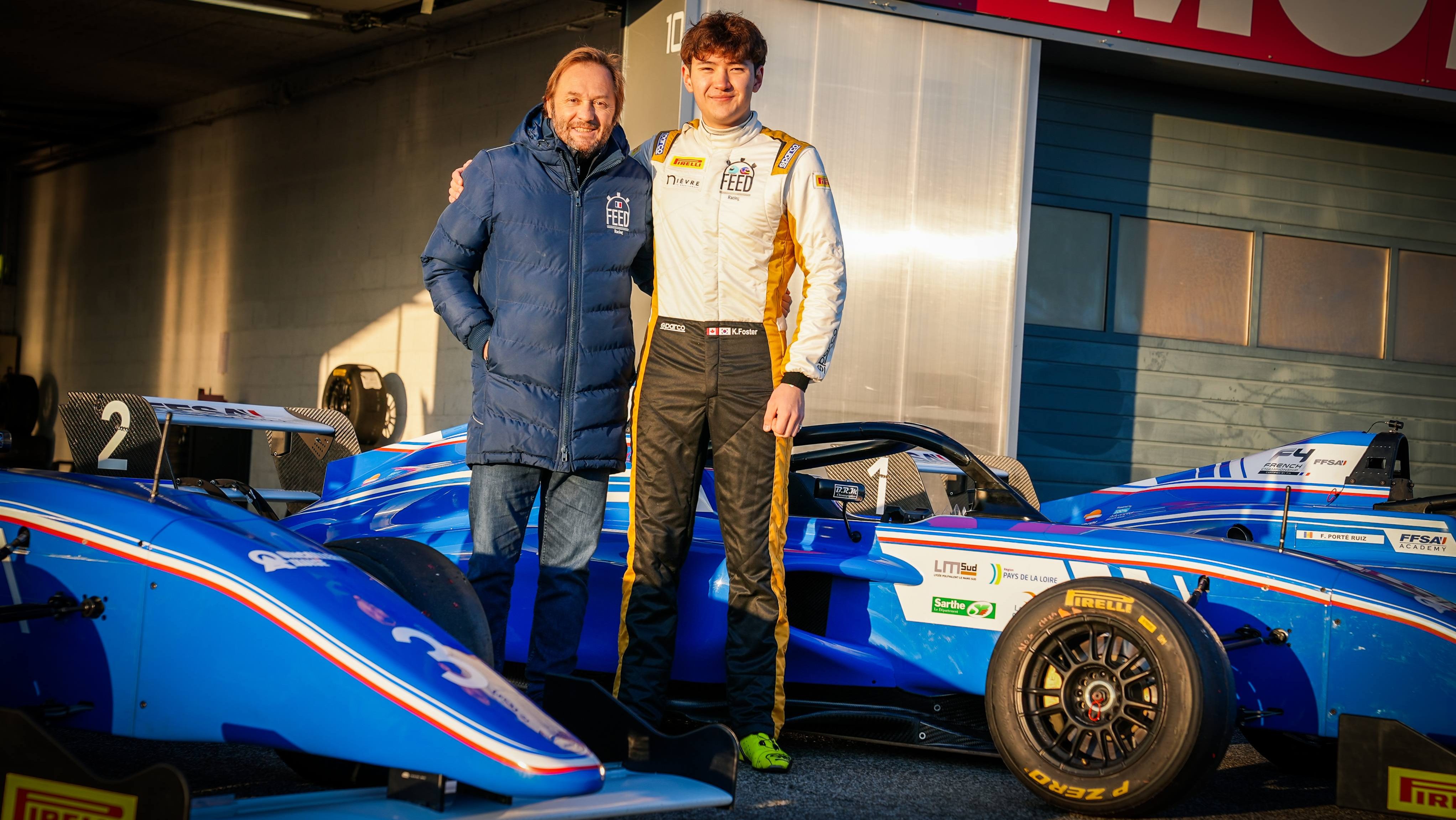 Patrick Lemarié, wearing a FEED Racing coat, puts his arm around Kevin Foster, wearing FEED Racing overalls. They stand amongst blue French F4 single-seaters in front of a garage.