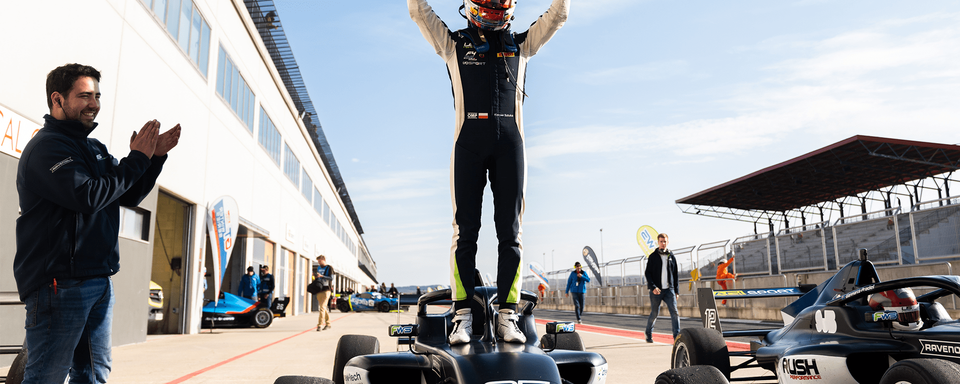 A driver stands on a black car facing the camera with his fists raised
