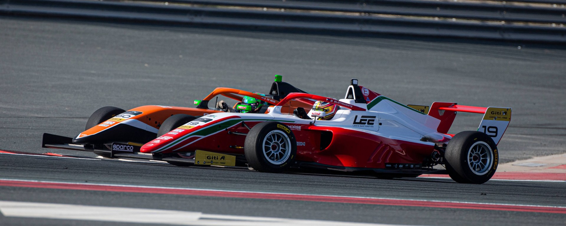 An orange and black car with a driver in a green helmet fighting a red and black car with a driver in a yellow and black helmet. The orange car is attempting an overtake on the inside of the corner.