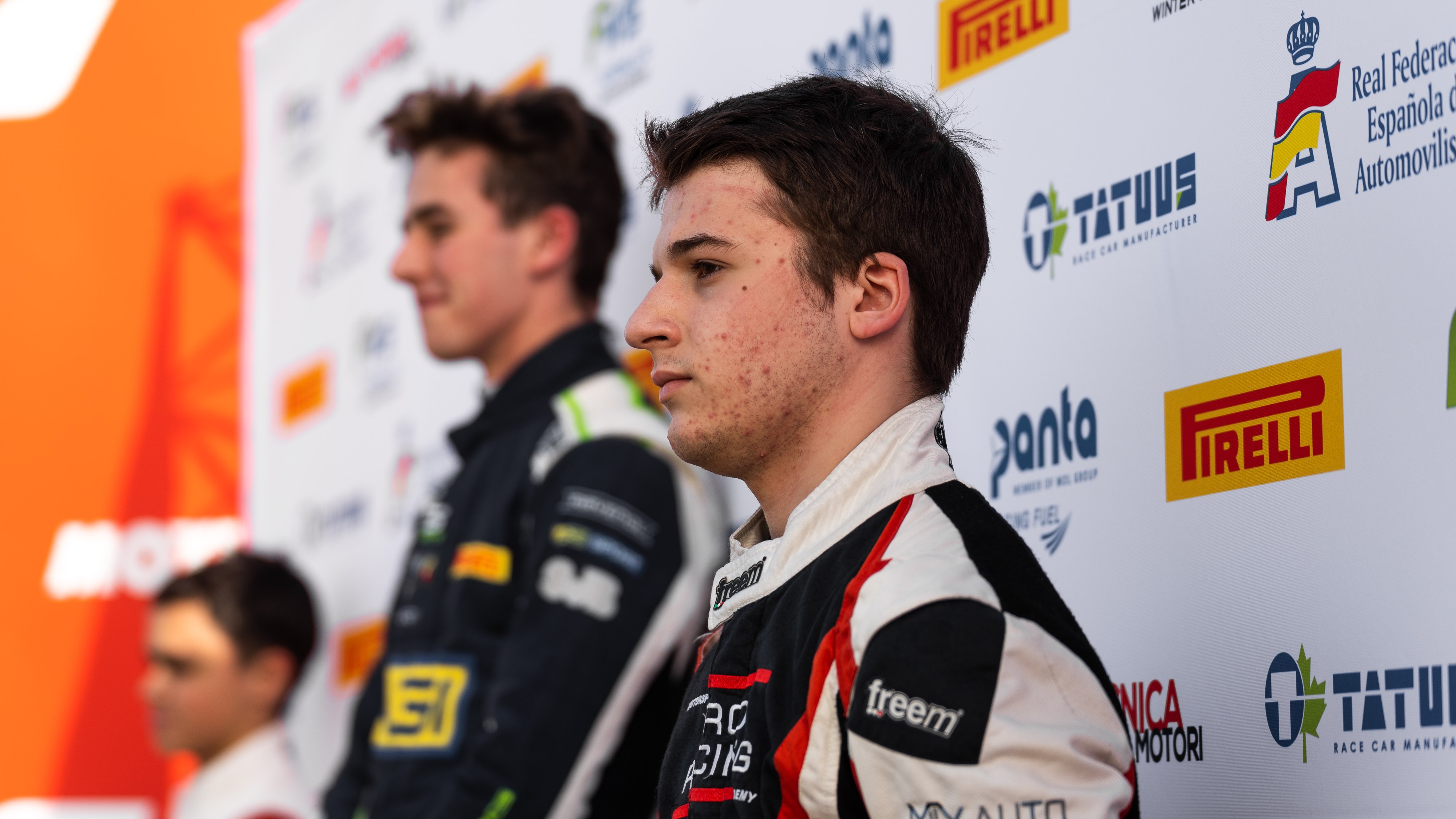 A driver stands on the third step of the podium looking to the left, with two other drivers in the background