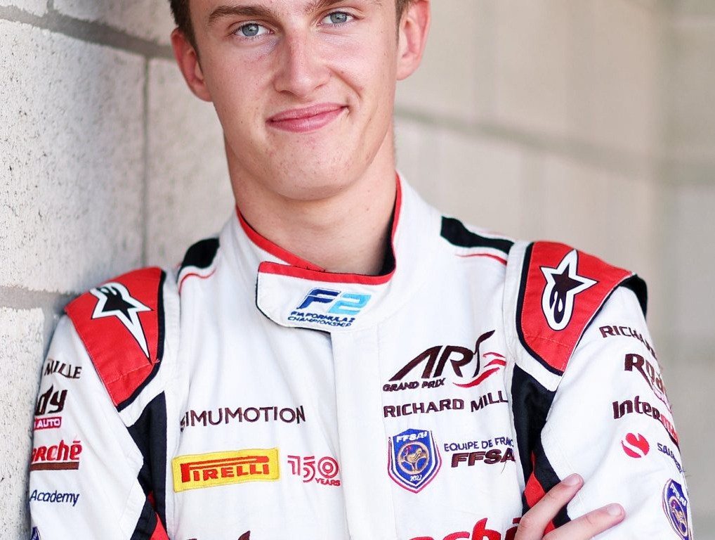Théo Pourchaire stands against a yellow brick wall with a red door in the background