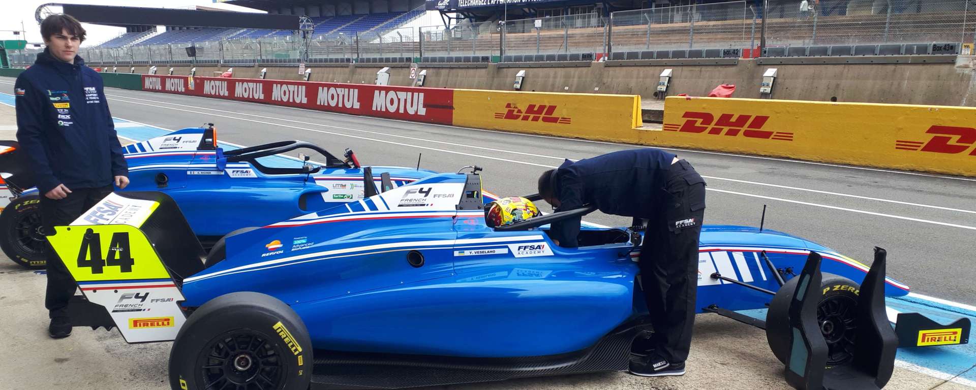 A blue and white car in pit lane, with a driver in the cockpit and an engineer leaning over to tighten belts