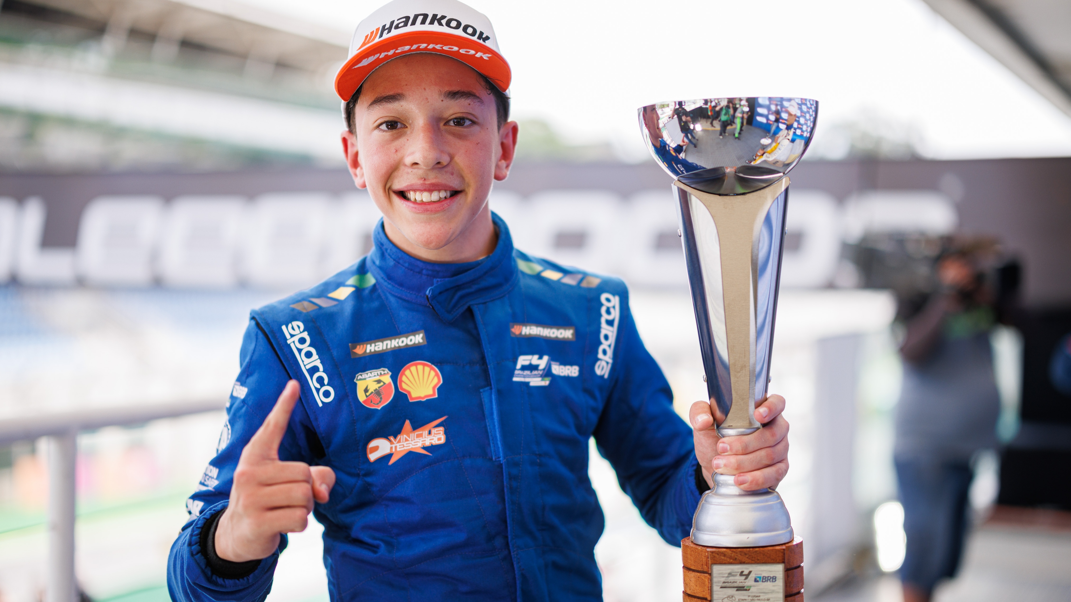A driver raises his finger and holds up a trophy while smiling at the camera