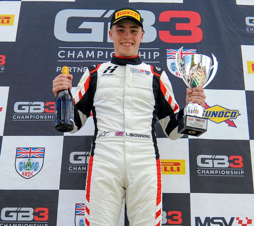 Luke Browning holds a trophy from Race 2 of the 2022 Snetterton 300 and a bubbly drink and smiles at the camera, pointed up at him