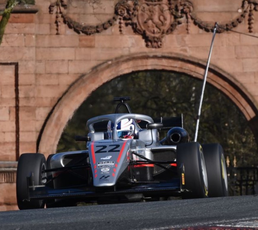 A silver car on a racetrack with an old bridge behind