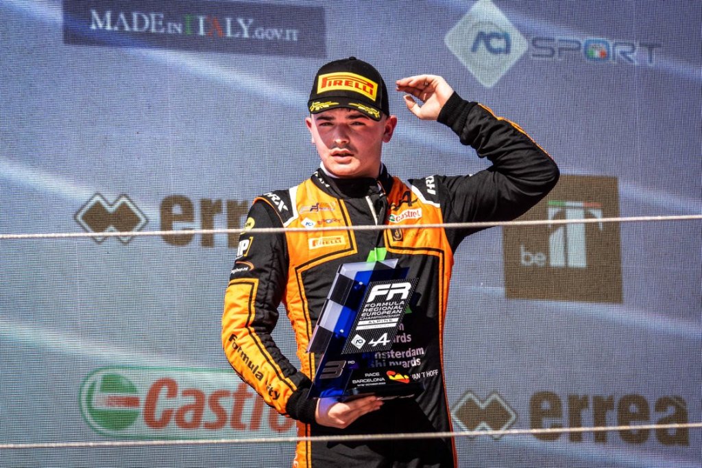 A racing driver wearing a black and orange race suit stands on a podium with a third place Formula Regional European Championship trophy