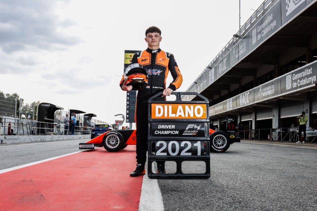 A racing driver wearing a black and orange race suit holds his orange helmet and a pit board that reads "Dilano: Spanish F4 Championship Driver Champion 2021"