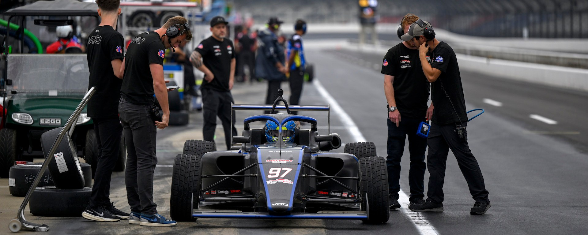 A blue and black car in pit lane with engineers beside and around it