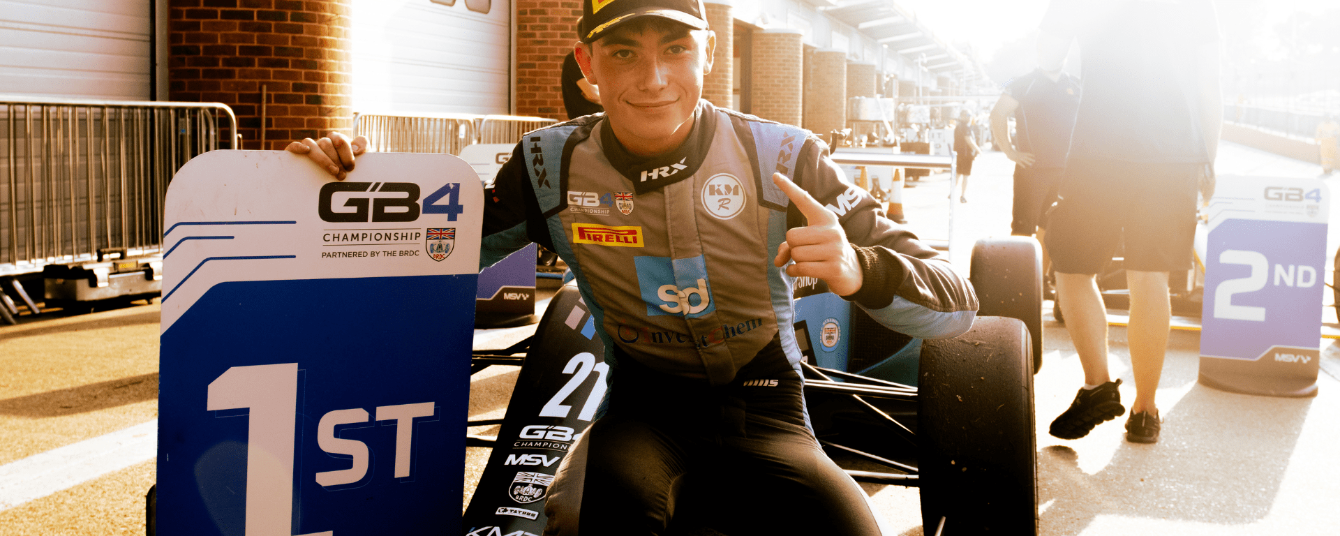 Tom Mills, the 2023 GB4 Champion, crouches in front of his car with a pointer finger raised and his other hand holding a first-place sign.