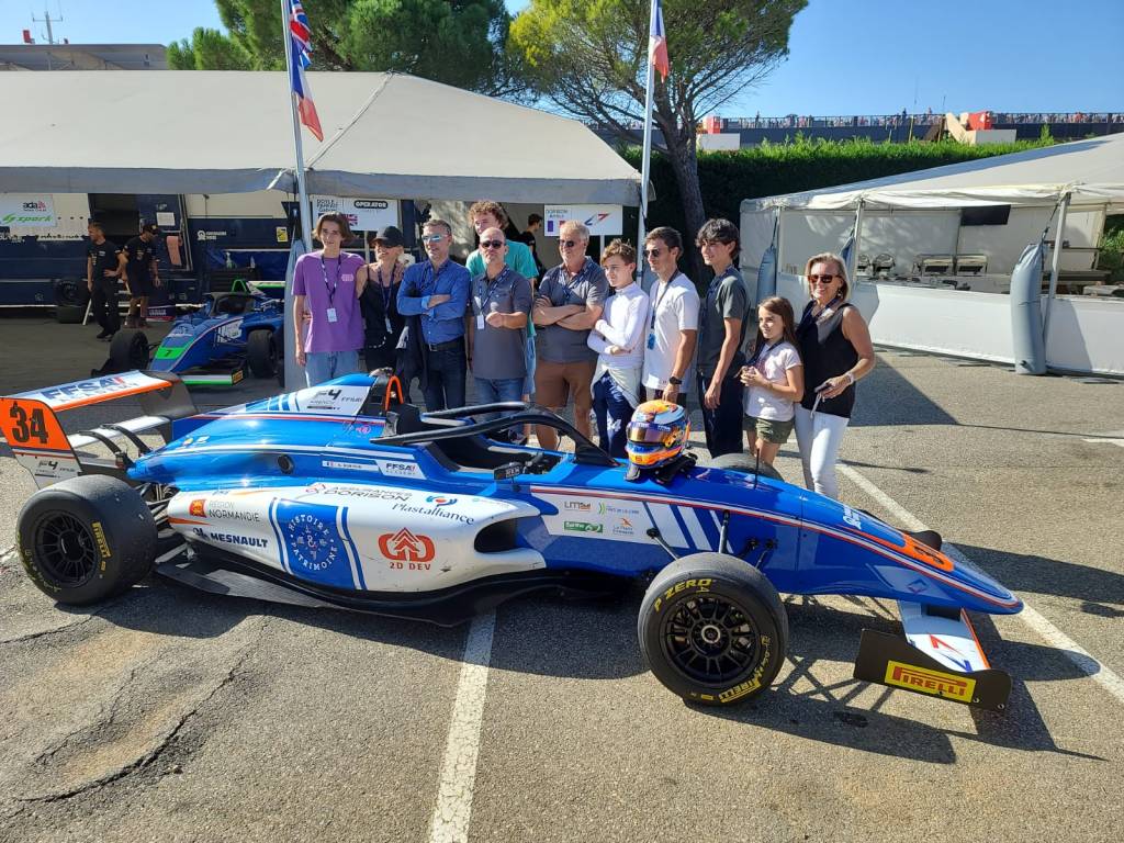 A group of 11 people of various ages standing behind an unoccupied blue racecar photographed from the right