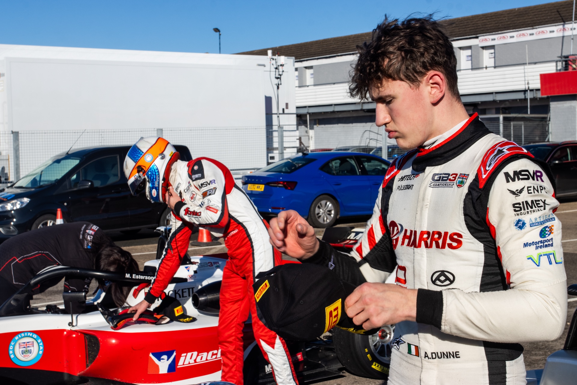 A driver in a white suit puts on a Pirelli hat while a driver in an iRacing livery steps out of his car