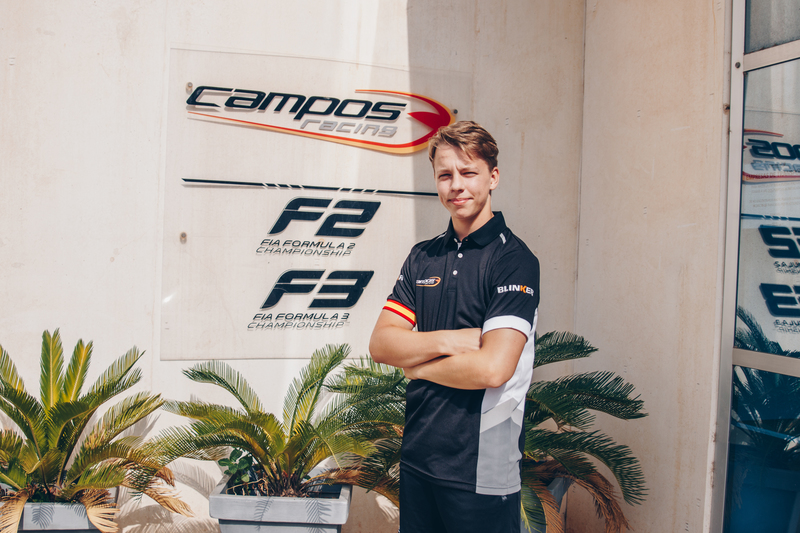 A young man in a black polo shirt in front of Campos Racing F2 and F3 logos