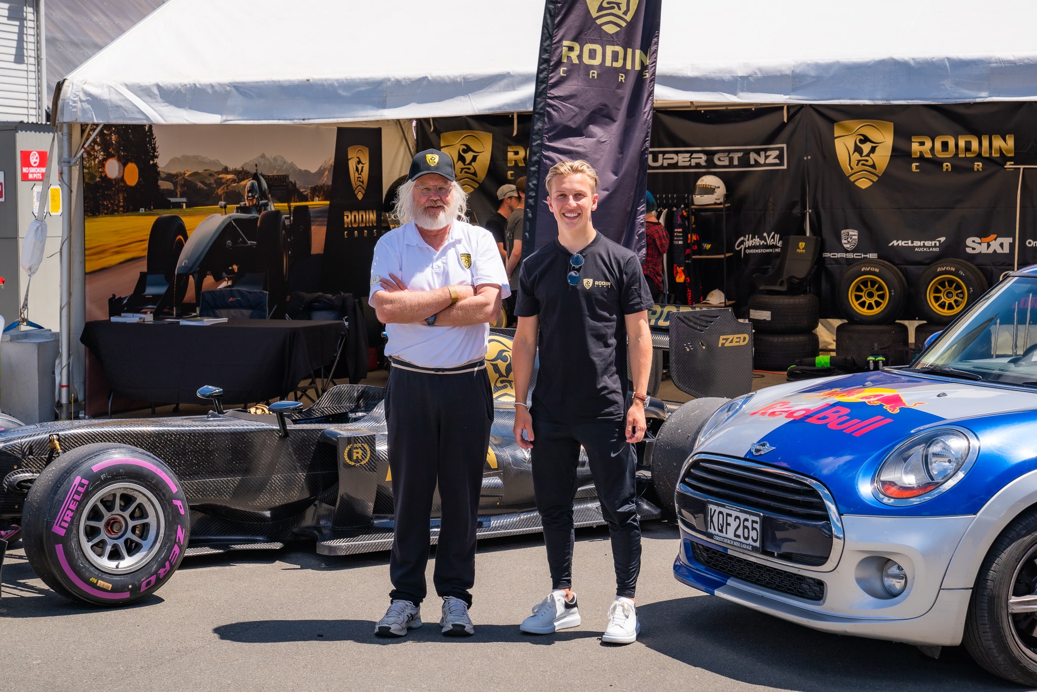 An older gentleman and a young man stand beside each other in front of a single-seater car and next to a Red Bull Mini Cooper